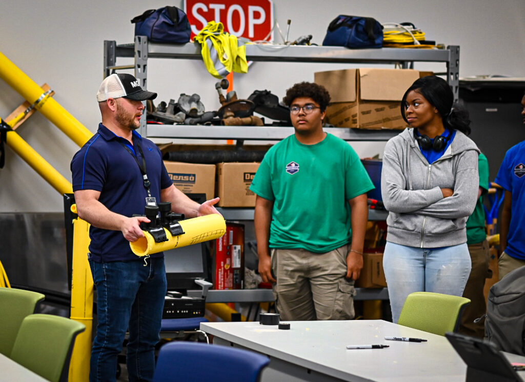 Ryan, Training guide, shows two students a piece of pipe and explains how it is used.