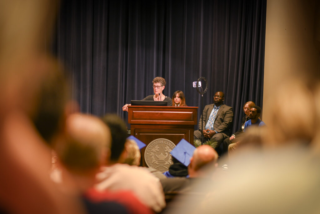 A woman stands at a podium speaking, she is framed by the heads of audience members