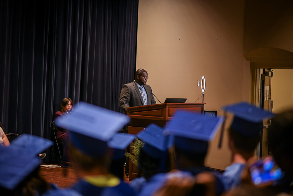 A man stands in the distance at a podium, graduation hats frame the image.