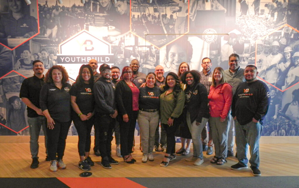 A group of people stand together in front of a big YouthBuild display.