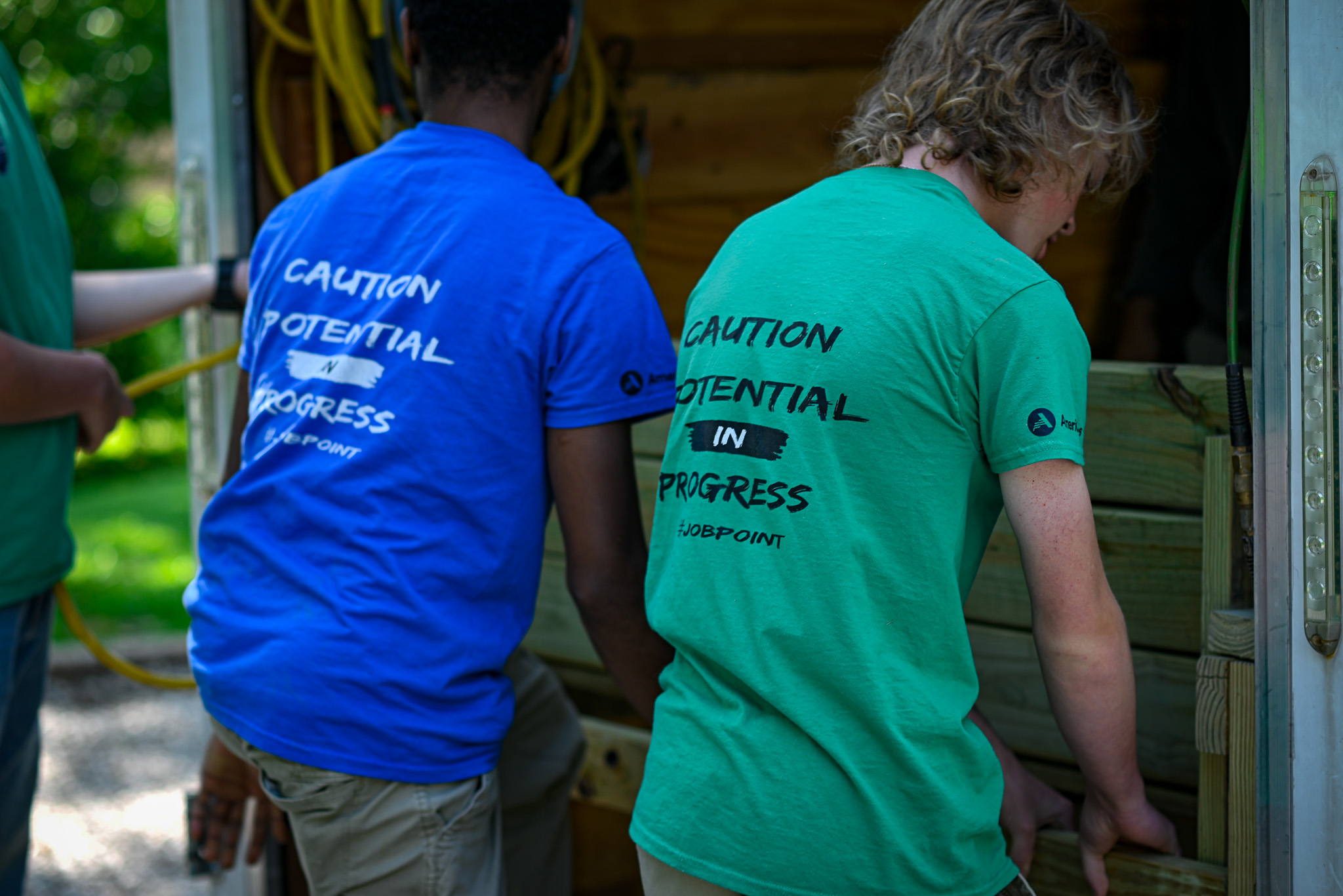 Two young men work together to unload a wooden bench from a covered trailer.