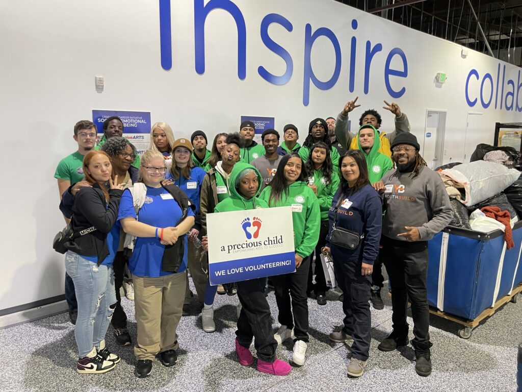 A group of young adults pose infront of a wall that says "Inspire" holding a sign that says "Precious child" 