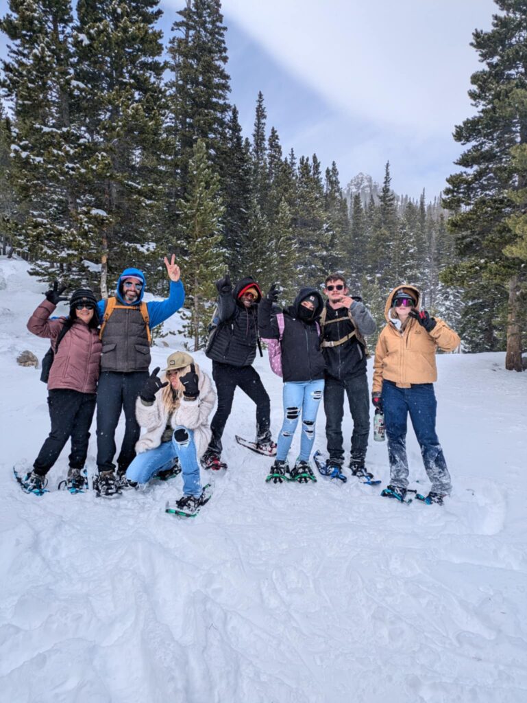 A group of people bundled in winter coats stand together on a snowy path between very tall pine trees. They have snow shoes on.