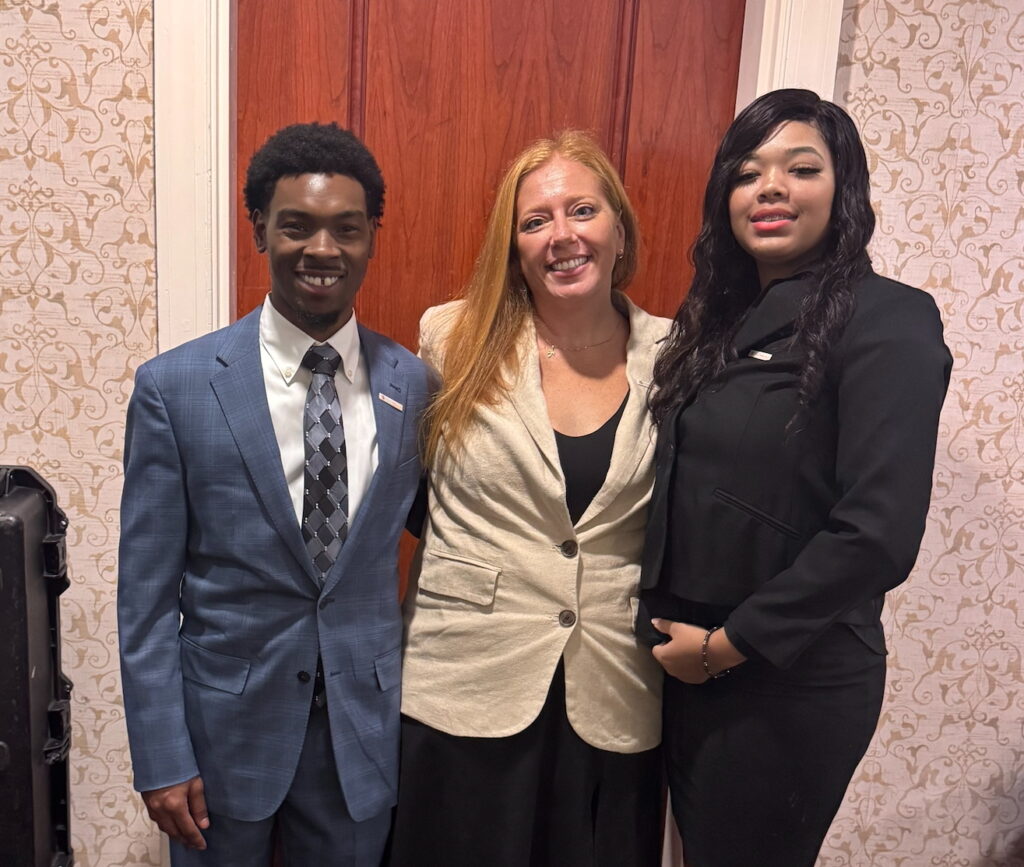 Two young people stand on either side of an adult leader in front of a brown door.